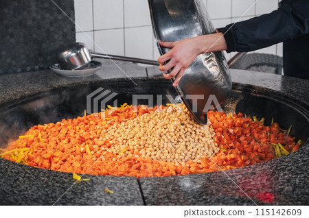 male chef adds chickpeas to cauldron with carrots to cook traditional oriental Uzbek pilaf in kitchen at restaurant in Uzbekistan male chef adds chickpeas to cauldron with carrots to cook traditional oriental Uzbek pilaf in kitchen at restaurant in Uzbekistan 115142609