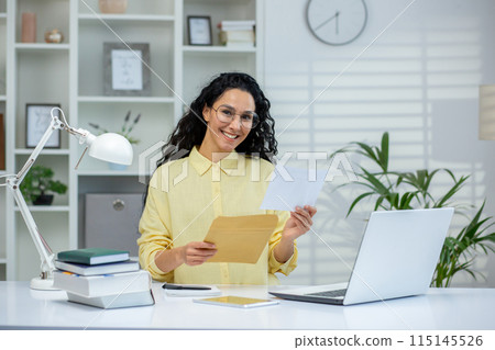 Smiling woman holding an envelope and letter while standing in a bright home office. Happy moment as she reads good news or a positive notification. 115145526