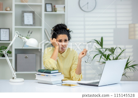 Woman looking stressed while sitting at a desk covered with books and a laptop in an office environment. Holding eyeglasses and resting head on hand. Woman looking stressed while sitting at a desk covered with books and a laptop in an office environment. Holding eyeglasses and resting head on hand. 115145528