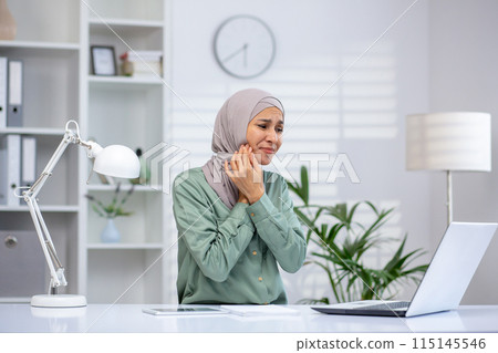 Woman wearing hijab experiencing toothache while working on laptop in a modern office. Concept of health issues at workplace. Woman wearing hijab experiencing toothache while working on laptop in a modern office. Concept of health issues at workplace. 115145546