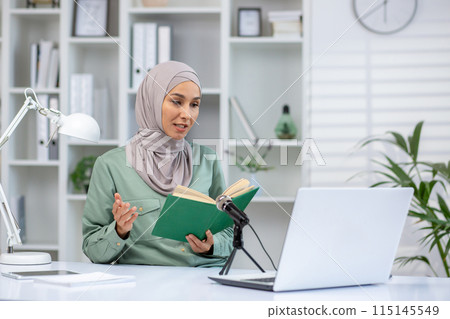 Woman in hijab recording a podcast while reading a book, sitting at a home office desk with laptop and microphone. Woman in hijab recording a podcast while reading a book, sitting at a home office desk with laptop and microphone. 115145549
