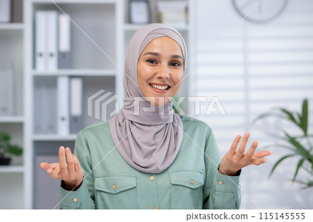 Smiling woman wearing hijab making a video call while working in a modern office setting with shelves and plants. 115145555