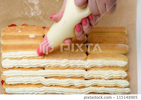 Women's hands cover rows of savoiardi with cream using a pastry bag. 115148905