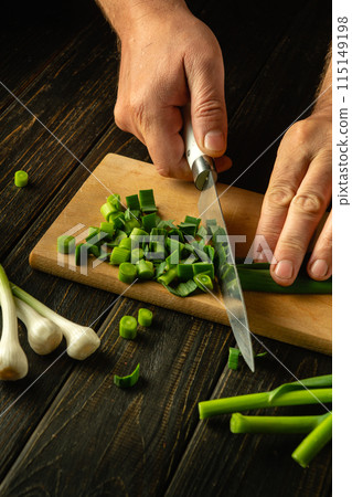 A cook cuts green garlic on a cutting board with a knife to prepare a vegetarian dish. Place for advertising 115149198
