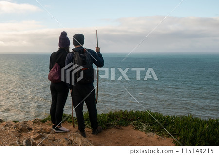 A young couple of tourists stands on a cliff in front of the Atlantic Ocean, a man and woman looking at the sea, hiking in the wild nature 115149215