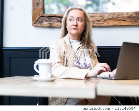 Positive woman using laptop and drinking coffee in cafe Positive woman using laptop and drinking coffee in cafe 115149594