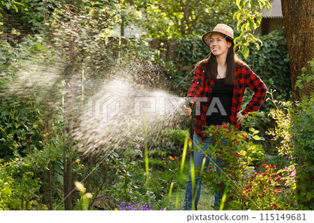 smiling young woman gardener watering flowers and plants in garden with hose smiling young woman gardener watering flowers and plants in garden with hose 115149681
