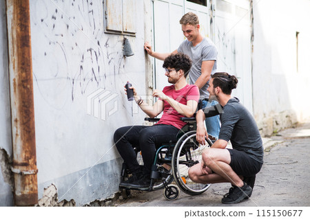 Young man in wheelchair spending free time outdoor with friends, drawin on abadoned building with spray paint. Male friendship. 115150677