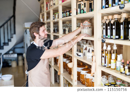 Handsome man working in package-free store using reusable containers. Zero waste shops offering package-free bulk goods and sustainable alternatives. 115150756
