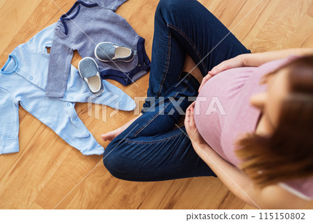 Pregnant woman sitting on floor touching her belly, looking at baby clothes. Pregnant woman sitting on floor touching her belly, looking at baby clothes. 115150802