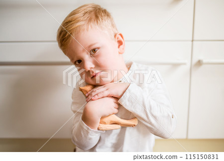 Portrait of a cute boy sitting on the floor in the kitchen, holding wooden horse toy. 115150831