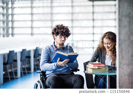 Handsome university student in wheelchair studying in library with classmate, preparing for final exam, presentation. Study group. Teamwork skills. 115150846