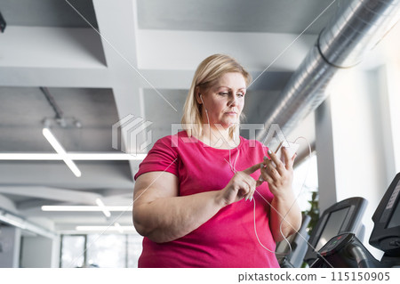 Overweight woman exercising on treadmill in gym, playing music on smartphone. 115150905