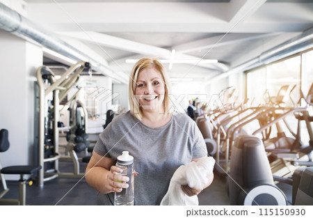 Overweight woman resting after workout in gym. Drinking water from bottle. Overweight woman resting after workout in gym. Drinking water from bottle. 115150930