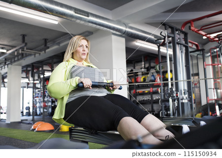 Overweight woman exercising on rowing machine. Routine workout for physical and mental health Overweight woman exercising on rowing machine. Routine workout for physical and mental health 115150934