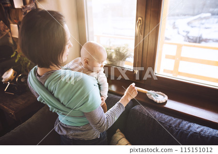 Mother holding small baby while dusting therir home, carrying him around in baby wrap. Household chores. 115151012