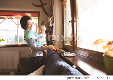 Mother holding small baby while dusting therir home, carrying him around in baby wrap. Household chores. 115151015