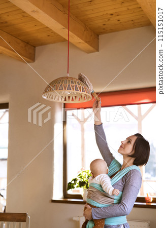 Mother holding small baby while dusting therir home, carrying him around in baby wrap. Household chores. Mother holding small baby while dusting therir home, carrying him around in baby wrap. Household chores. 115151017