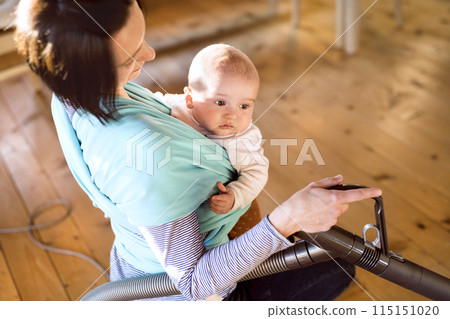 Mother holding small baby while vacuuming floor, carrying him around house in baby wrap. Household chores. Mother holding small baby while vacuuming floor, carrying him around house in baby wrap. Household chores. 115151020