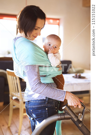 Mother holding small baby while vacuuming floor, carrying him around house in baby wrap. Household chores. Mother holding small baby while vacuuming floor, carrying him around house in baby wrap. Household chores. 115151022