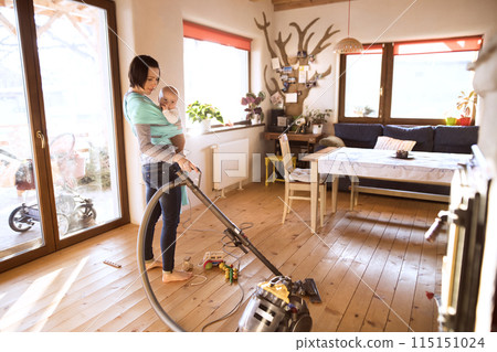Mother holding small baby while vacuuming floor, carrying him around house in baby wrap. Household chores. 115151024