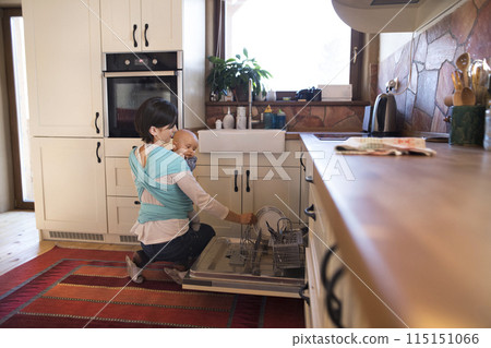 Mother holding small baby while loading dishwasher, carrying him around house in baby wrap. Household chores. 115151066