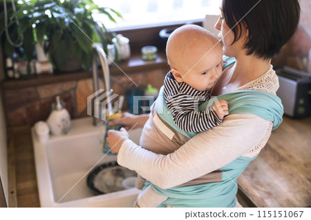 Mother holding small baby while cleaning dishes, carrying him around house in baby wrap. Household chores. Mother holding small baby while cleaning dishes, carrying him around house in baby wrap. Household chores. 115151067