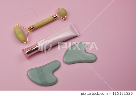Gua Sha face tools, green stone scrapers, a massage roller and a tube of cream on a pink background, spa, skin care Gua Sha face tools, green stone scrapers, a massage roller and a tube of cream on a pink background, spa, skin care 115151526