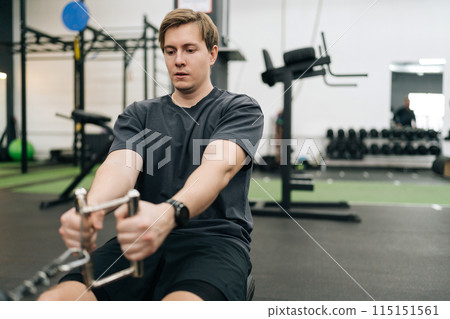 Closeup of focused motivated sportsman working out on stationary rowing machine in gym. Young beginner sporty man doing exercises on fitness machine at healthy club. Concept of healthy sports life. Closeup of focused motivated sportsman working out on stationary rowing machine in gym. Young beginner sporty man doing exercises on fitness machine at healthy club. Concept of healthy sports life. 115151561