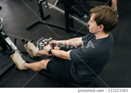 Rear top view of dedicated sportsman gym workout on rowing machine for wellness and strong body with focus for goals. Beginner athletic male training for muscle development, motivation and cardio. Rear top view of dedicated sportsman gym workout on rowing machine for wellness and strong body with focus for goals. Beginner athletic male training for muscle development, motivation and cardio. 115151563