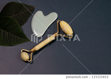Jade face roller and gua sha massage tool on green leaf. Gua Sha face tool, green stone massage roller on a dark background with copy space, spa background with place for text, skin care Jade face roller and gua sha massage tool on green leaf. Gua Sha face tool, green stone massage roller on a dark background with copy space, spa background with place for text, skin care 115151653