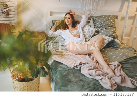No retouch portrait of a young natural beauty woman at home bedroom cozy rustic interior at vacation countryside house. Beautiful slim sexy lady in white male shirt and lace lingerie lying in bed 115153157