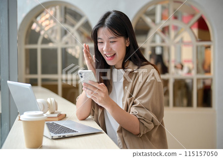 A cheerful Asian woman is sitting in a cafe and looking at her smartphone with an excited expression. A cheerful Asian woman is sitting in a cafe and looking at her smartphone with an excited expression. 115157050