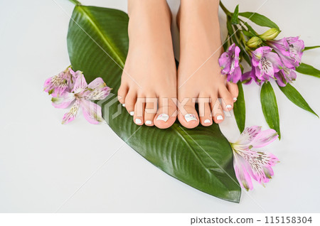 Beautiful perfect female skin legs feet top view with tropical flowers and green palm leaf. Nail polish, care and clean, spa pedicure treatment in white. Concept on background isolated. Copy space Beautiful perfect female skin legs feet top view with tropical flowers and green palm leaf. Nail polish, care and clean, spa pedicure treatment in white. Concept on background isolated. Copy space 115158304