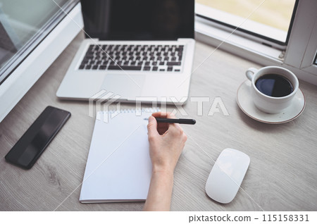 Unrecognizable woman hands writing in empty mock up notebook and computer. Freelancer using smart phone and laptop at home during quarantine. 115158331