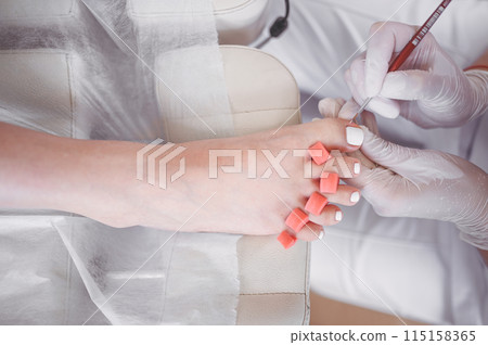 Pedicurist doing white nail polish on client legs using shellac lamp. Professional medical pedicure procedure. Foot treatment in SPA salon. Podiatry clinic. Beautician doctor hands in white gloves. 115158365