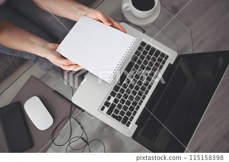 Unrecognizable woman hands writing in empty mock up notebook and computer. Freelancer using smart phone and laptop at home during quarantine. 115158398