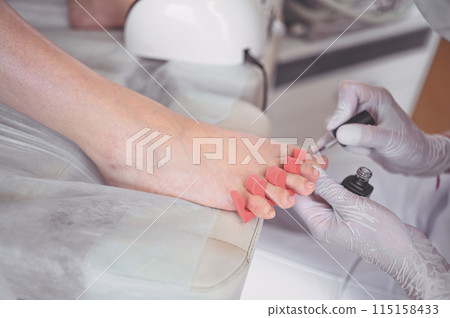 Pedicurist doing white nail polish on client legs using shellac lamp. Professional medical pedicure procedure. Foot treatment in SPA salon. Podiatry clinic. Beautician doctor hands in white gloves. 115158433