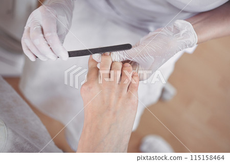 Pedicurist doing professional medical pedicure procedure in beauty salon using nail file. Foot treatment in SPA salon. Podiatry clinic. Beautician doctor hands in white gloves with female legs 115158464