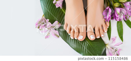 Beautiful perfect female skin legs feet top view with tropical flowers and green palm leaf. Nail polish, care and clean, spa pedicure treatment in white. Concept on background isolated. Copy space 115158486