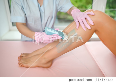 Woman with long tanned perfect legs and smooth skin having wax stripe depilation hair removal procedure on legs in beauty salon. Beautician in blue robe, pink gloves. Body care, epilation spa concept Woman with long tanned perfect legs and smooth skin having wax stripe depilation hair removal procedure on legs in beauty salon. Beautician in blue robe, pink gloves. Body care, epilation spa concept 115158488