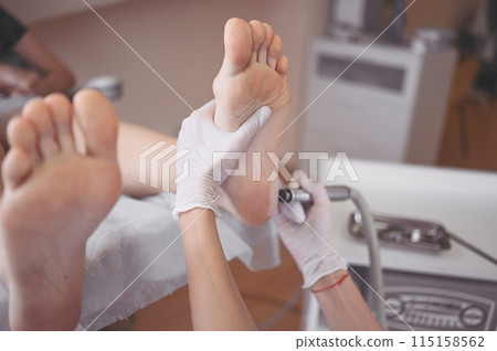Podiatrist using grinding equipment and making procedure polish for feet pedicure. Podology beautician in white gloves cleaning skin of client legs from callus and corn with professional tool. 115158562