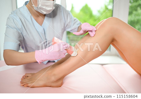 Woman with long tanned perfect legs and smooth skin having wax stripe depilation hair removal procedure on legs in beauty salon. Beautician in blue robe, pink gloves. Body care, epilation spa concept Woman with long tanned perfect legs and smooth skin having wax stripe depilation hair removal procedure on legs in beauty salon. Beautician in blue robe, pink gloves. Body care, epilation spa concept 115158604