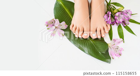 Beautiful perfect female skin legs feet top view with tropical flowers and green palm leaf. Nail polish, care and clean, spa pedicure treatment in white. Concept on background isolated. Copy space 115158655