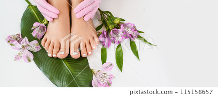 Beautiful perfect skin female legs feet top view with tropical flowers and green palm leaf. Hands of beautician in pink gloves. Nail polish, care and clean, spa pedicure treatment in white. Copy space 115158657