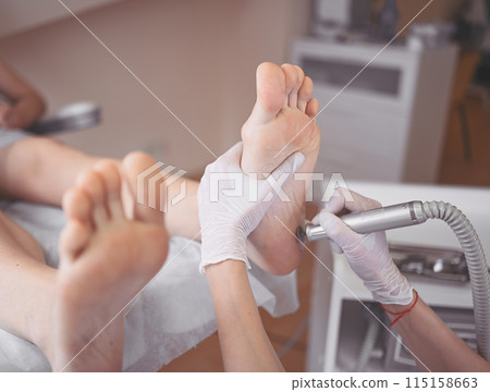 Podiatrist using grinding equipment and making procedure polish for feet pedicure. Podology beautician in white gloves cleaning skin of client legs from callus and corn with professional tool. 115158663