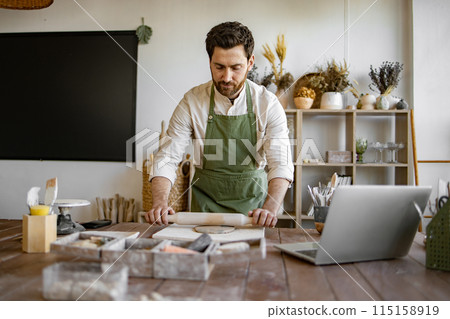 Mature male potter flattens clay with rolling pin to create handmade dishes. Mature male potter flattens clay with rolling pin to create handmade dishes. 115158919