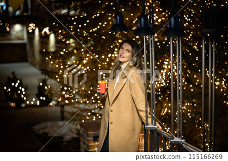 beautiful young girl in a beige coat with a paper glass of coffee against the backdrop of many bokeh lights beautiful young girl in a beige coat with a paper glass of coffee against the backdrop of many bokeh lights 115166269