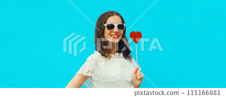 Portrait of beautiful young woman with red heart shaped lollipop on stick on blue studio background Portrait of beautiful young woman with red heart shaped lollipop on stick on blue studio background 115166881