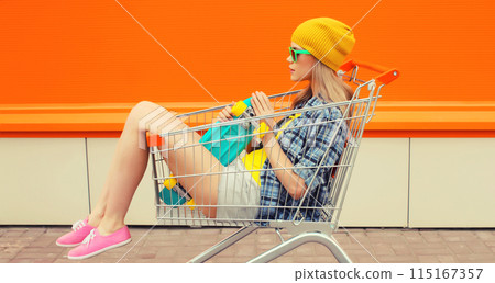 Stylish modern young woman sitting in shopping trolley cart in colorful clothes on city street 115167357
